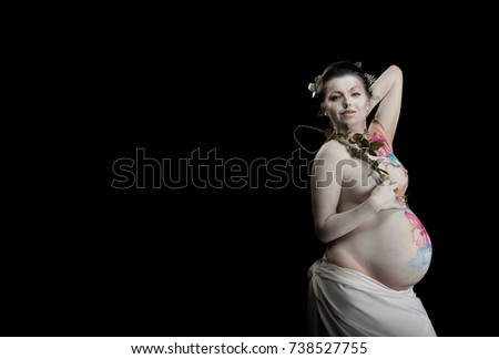 Emotional actress, pregnant woman with bodypainting on body and flowers orchid in hands posing on black background. White leather with large flowers and colored ornaments.