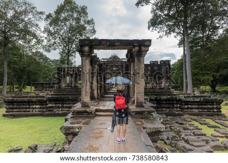 Young female with red backpack and map,raise an umbrella at the ancient Prasat Baphuon in a sprinkle of rain,Khmer temple ,Siem Reap,Cambodia - traveling,take a photos concept.