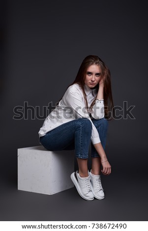 Cute European teenage lady sit on wooden box in blue jeans and white classic shirt