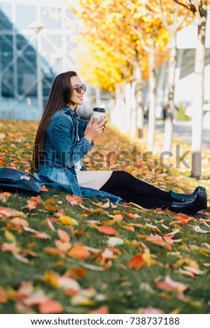Bright picture of young fashion woman drinking coffee from disposable cup and sitting on the grass strewed with yellow and red leaves. Education, lifestyle and people concept.