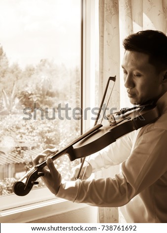 Young Asian man playing violin at window. Classical music instrument. String equipment. Art and music portrait background. Light and shadow contrast. Sepia color tone.