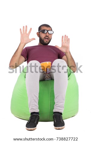 Handsome beard young man with a cinema glasses sitting on a green chair and watching a film, guy wearing red t-shirt and gray pants, isolated on white background
