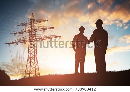 Silhouette Of Two Engineers Shaking Hands With Electricity Pylon Against Dramatic Sky Royalty-Free Stock Photo #738900412
