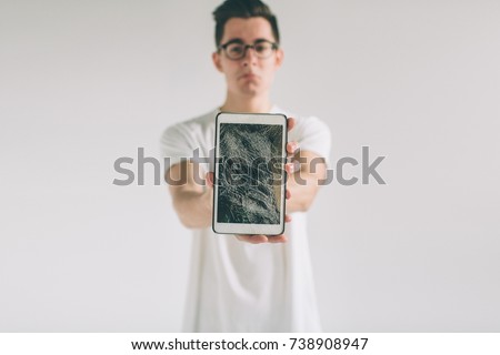 Nerd is wearing glasses. Student presenting a broken black tablet behind glass. upset man holds a out-of-use tablet or smartphone. Isolated on a light background. Broken screen