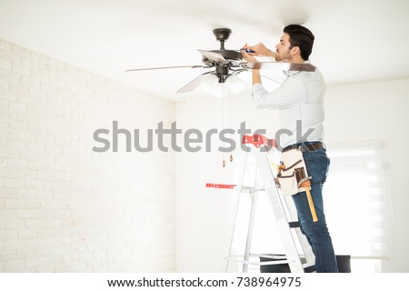 Profile view of a male electrician stepping on a ladder and installing a ceiling fan in a house Royalty-Free Stock Photo #738964975