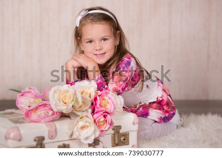 Cute baby girl with brunette hairs pink cheeks and green eyes posing sit for camera in studio wearing airy colorful dress and fancy casuals pumps close to traveling trunk bag with pictures and roses