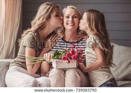 Beautiful senior woman is sitting with presents and smiling while her daughter and granddaughter are kissing her in cheeks