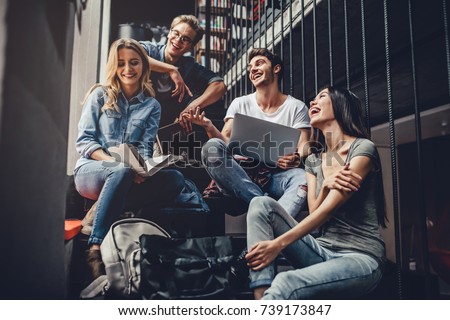 Students are studying in library. Young people are spending time together. Reading book, working with laptop and communicating while sitting on stairs in library. Royalty-Free Stock Photo #739173847