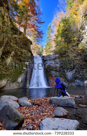 Happy kid near photo camera on tripod with a waterfall in background in autumn