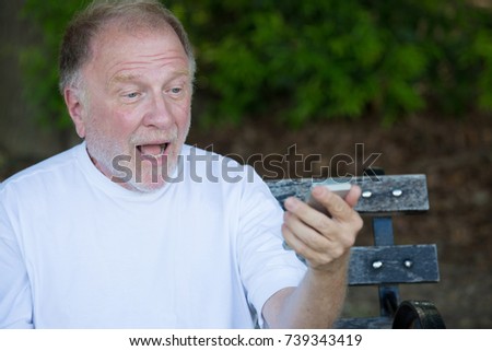 Closeup portrait of bold elderly man in white shirt, shocked at checking smartphone, sending text message, seated on a bench, isolated outdoor background.