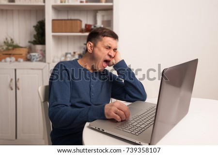 Caucasian middle-aged man is sitting at the table and yawns in front of laptop in the dining room