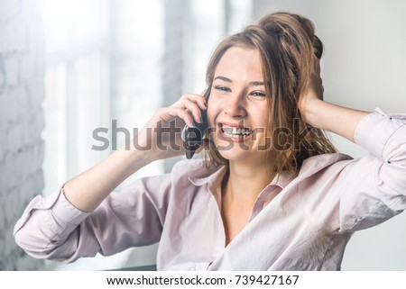Pretty young smiling brunette girl talking by smartphone sitting on black modern chair, indoor white bricks wall
