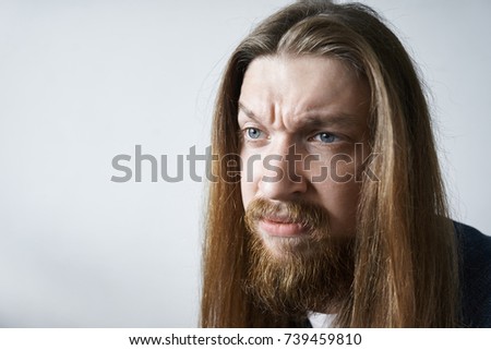 Negative human emotions. Displeased fed up young European bearded male with long loose hair frowning eyebrows and pouting lips feeling unwillingness towards doing something boring or unpleasant