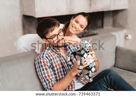The couple plays with the robot a rhinoceros. The guy sits on the couch and holds the robot in his hands. The girl is standing behind him. She hugs the guy and looks at the robot.
