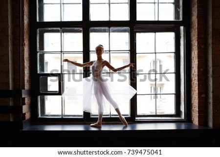 Full length picture of female classical ballet dancer in white tutu dancing in ballet studio, standing on wide windowsill and looking at camera. People, profession, hobby and occupation concept