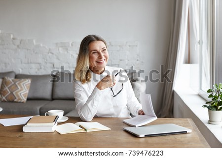 Indoor portrait of cheerful middle aged female chief editor with thick loose hair sitting at her workplace and laughing while reading satirical verses, holding book, enjoying author's writing style Royalty-Free Stock Photo #739476223