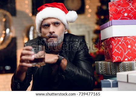 Bad Santa Claus in a black leather jacket sits with a cigar in his mouth and a glass of whiskey in his hands surrounded by gifts. and the background is a poorly decorated place for a holiday.