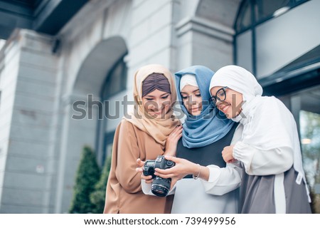 Traveling and photography. Young muslim women in beautiful hijab holding the camera and looking into it. They are smiling. Outdoors in town.