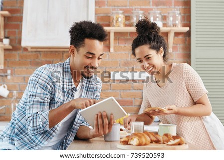 Cheerful couple choose new furniture in kitchen, look happily into screen of tablet. Beautiful curly wife makes sandwiches, being pleased to watch funny pictures or video on electronic device