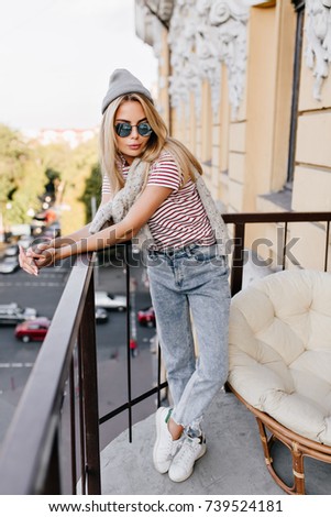 Full-length portrait of trendy girl in white sneakers standing with legs crossed and looking with interest. Slim young lady wears jeans and sunglasses has photoshoot on terrace.