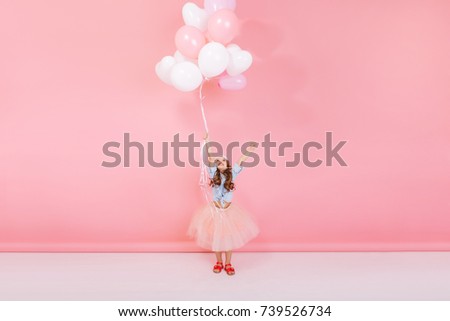 Happy brightful image of excited joyful little girl in tulle skirt holding ribbon with flying balloons isolated on pink background. Looking above, expressing happiness, joy at birthady party