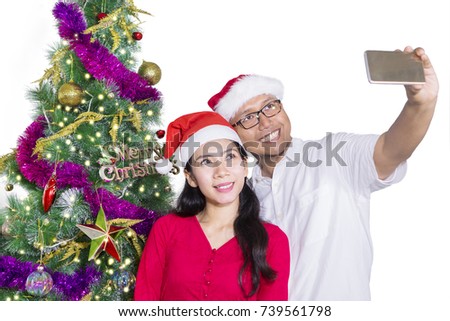 Happy couple celebrating Christmas day while wearing Santa hat and making selfie photo with Christmas tree background, isolated on white background