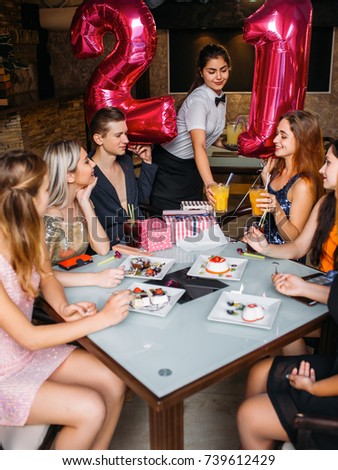 Joyful birthday party with friends in cafe. Smiling youth company with balloons, waitress with drinks. Modern leisure time, happy atmosphere, festivity concept