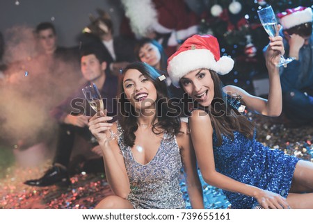 Two girls are sitting on the floor. Around them is scattered confetti. They rest after the party for the new year and laugh happily. They have glasses with champagne in their hands.