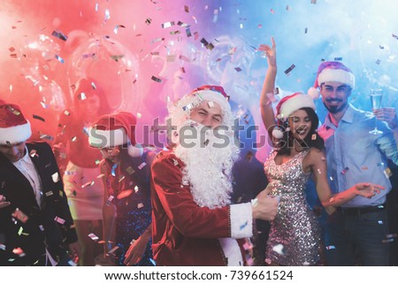 A man dressed as Santa Claus has fun at a New Year party. Together with him have fun friends. Around them flies confetti, against a background of multi-colored haze. People are very cheerful.