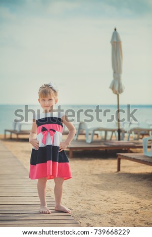Young cute baby girl enjoying childhood summer time on sandy beach posing on wooden pier bridge wearing casual stylish dress.
