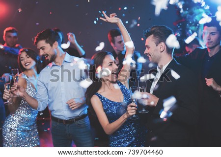 Young people have fun at a New Year's party. In the foreground two pairs dance. The girls are wearing gray and blue dresses in sequins. On one of the guys a light shirt, on the other a black jacket.