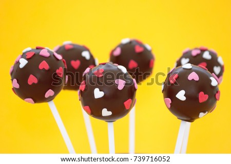 Chocolate cake pops, decorated with pink and white confectionery sprinkles in form of heart on a yellow background. Picture for a menu or a confectionery catalog.
