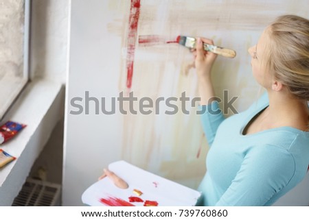 Young blonde female painter painting picture with brush in atelier