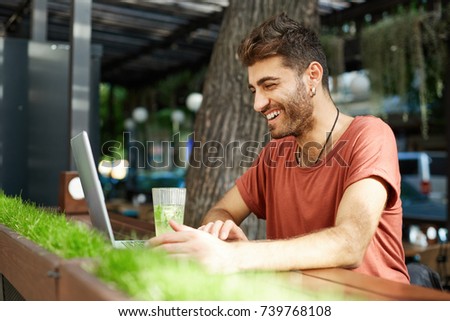 Glad bearded man casually dressed using laptop computer, smiling and laughing while talking with video with his girlfriend, drinking lemonade, sitting at wooden table at summer platform of cafeteria