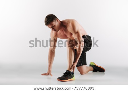 Picture of displeased young sportsman standing isolated over grey background. Looking aside have painful feelings in leg.