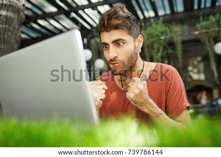 Shot of worrying focused man with stylish hairstyle dressed casually closely looking at the screen of his notebook, watching online translation of match, clenching his fists in nerves.