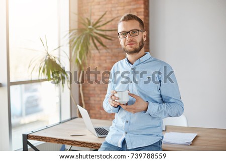 Close up portrait of young attractive company founder in glasses and casual outfit, standing in personal office, holding cup of coffee in hands, looking in camera with relaxed expression, posing for
