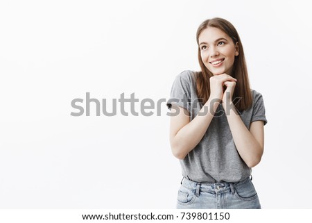 Close up of charming good-looking young student girl with dark hair and eyes in casual outfit smiling, looking aside with dreamy and happy face expression, holding hands together under face.