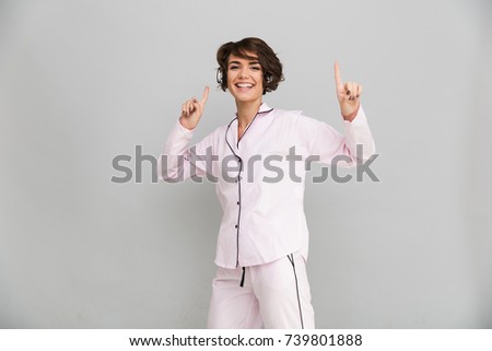 Portrait of a happy cheerful girl in pajamas listening to music with headphones and dancing isolated over gray background