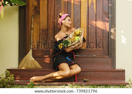 Beautiful woman with basket full of exotic fruits is sitting on the doorstep beside her house