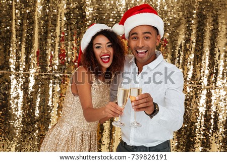 Portrait of smiling cheerful african couple in red hats celebrating New Year while toasting with champagne glasses isolated over golden shiny bakground
