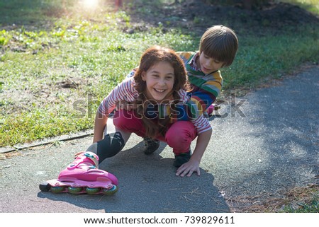 A little boy raises his sister who fell on roller skates. The boy helps the girl who fell, rolling on rollers in a park.