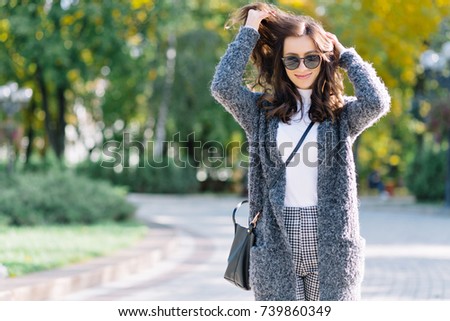 Style woman with charming smile and dark hair is walking in the autumn park in sunlight. She is posing at camera and has wonderful big blue eyes. Background park. Beautiful portrait.