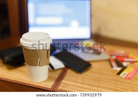 White paper Cup with burning coffee on the background of laptop and smartphone. Scattered Bank cards and a black purse. Work on the go.