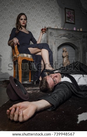Film scene. Femme Fatale committed murder. Beautiful woman sits near dead body of detective. Body of man lying on floor. Woman holding glass of whiskey