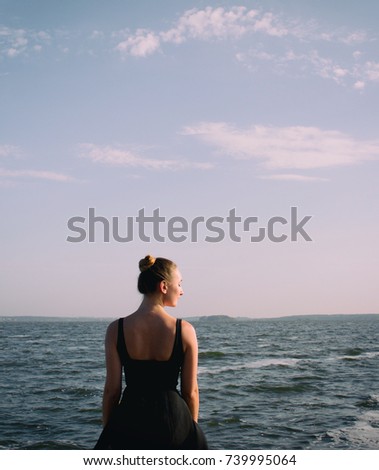 Beautiful girl looks at the sea. Young girl in a dress. Young blonde girl looking at a calm sea and blue skies back view. Retro styled photo.