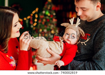 Parents play with little daughter in deer hat holding her on their arms and standing before a Christmas tree