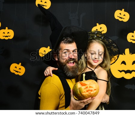 Halloween party concept. Girl and bearded man with cheerful faces on black background with decor. Wizard and little witch in black hats hold pumpkin and hug. Father and daughter in costumes
