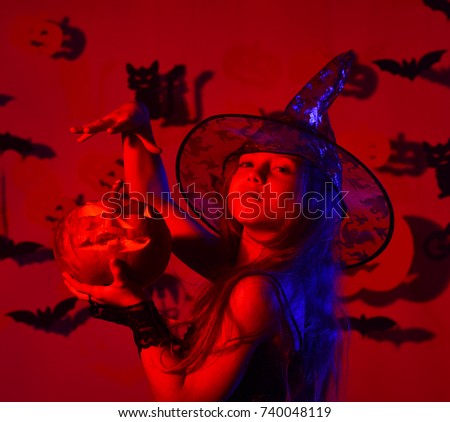Kid in spooky witches costume holds jack o lantern. Little witch wearing black hat. Halloween party and decorations concept. Girl with proud face on bloody red background with bats and pumpkins decor