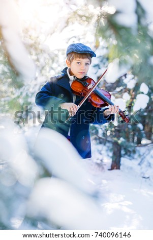 Winter fairy tale. Portrait of cute boy violinist in white winter forest. Playing classic instrument. Dressed in coat. Artistic looking.